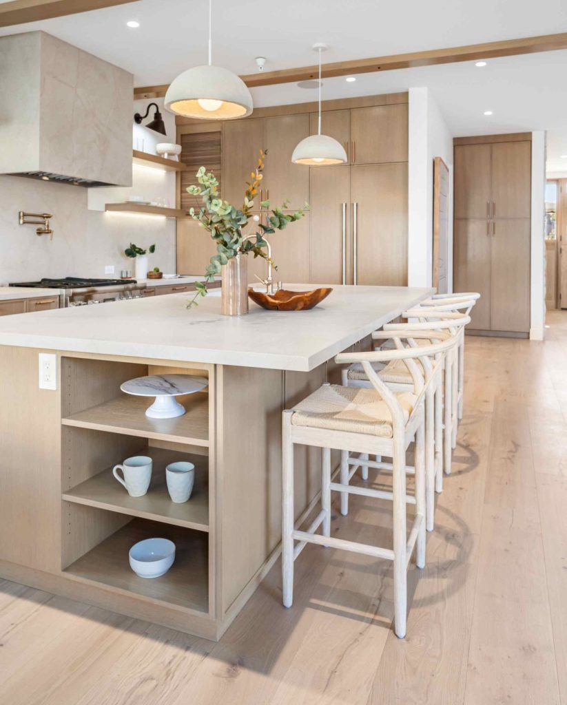 Modern kitchen with light wood cabinets, a large island featuring open shelves with dishes, three white barstools, pendant lights overhead, and minimal decorative accents, including a vase with flowers.