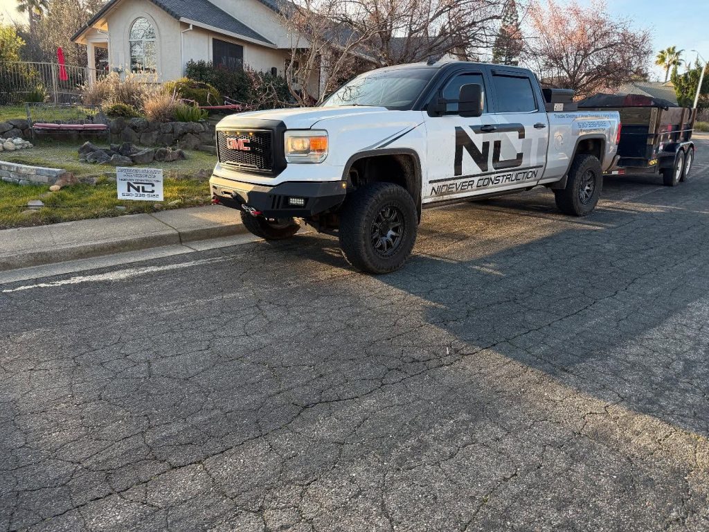 A white GMC pickup truck with "Nicewicz Construction" branding is parked on a residential street next to a yard sign with the same logo; a trailer is attached to the truck. A house and garden are visible in the background.