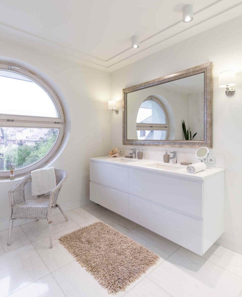 Bright, modern bathroom with a large round window, wicker chair, beige rug, white vanity with twin sinks, and a large framed mirror above. Soft lighting and minimal decor create an airy, clean atmosphere.