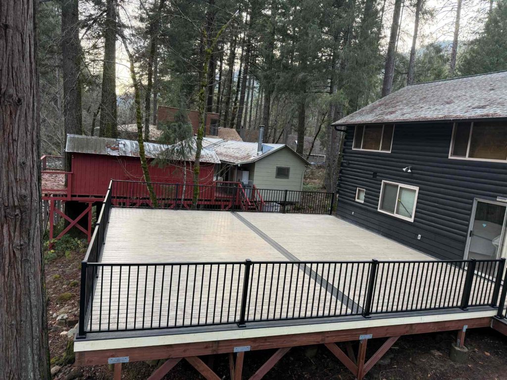 A large wooden deck with black railings is attached to a dark house, overlooking a sloped, forested area with tall trees and nearby red and gray buildings in the background.